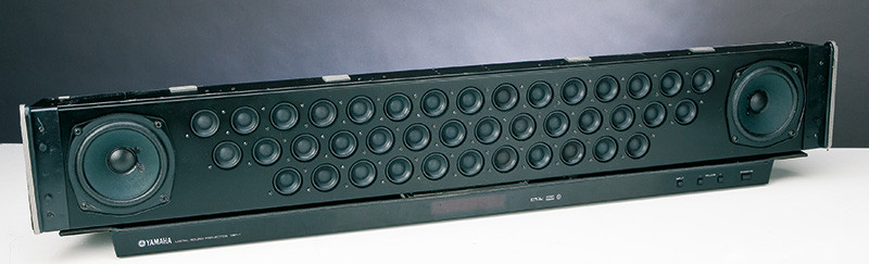 Il Digital Sound Processor Yamaha YSP-1 (in alto senza griglia di protezione), è il progenitore dei numerosi “Sound Bar” da mettere sotto al televisore per simulare la presenza nella stanza di un sistema di altoparlanti multicanale. Le sorgenti virtuali vengono simulate dirigendo un “raggio sonoro” contro le pareti in modo che raggiunga l’ascoltatore da un punto completamente diverso da quello (frontale) da cui è stato emesso. Il “trucco” consiste nell’inviare ai 40+2 altoparlanti (ognuno dotato del proprio amplificatore!) un segnale opportunamente ritardato in modo tale che la “linea di suono” generi un raggio molto ristretto ed orientato nella direzione opportuna. L’elaborazione, evidentemente non invariante, deve essere distinta per ciascuna delle sorgenti virtuali che si desidera simulare, salvo poi emettere da ciascun altoparlante la somma che gli compete. Qui sopra, l’YSP-800: presentato nel 2005 come fratellino minore, utilizzava invece “solo” 21+2 altoparlanti ed altrettanti amplificatori.